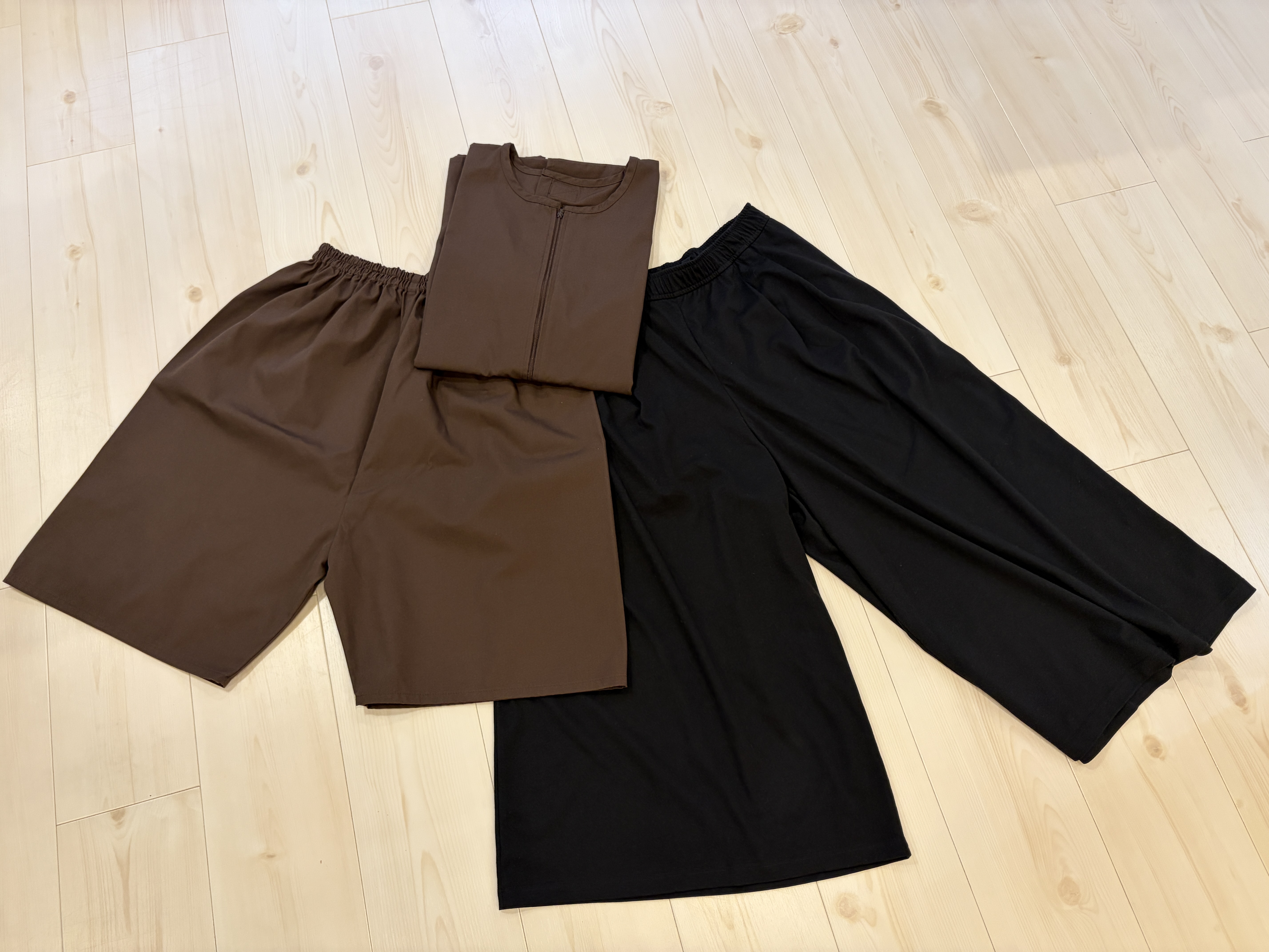 Brown and black clothing items laid out on light wooden floor including shorts, shirt, and pants