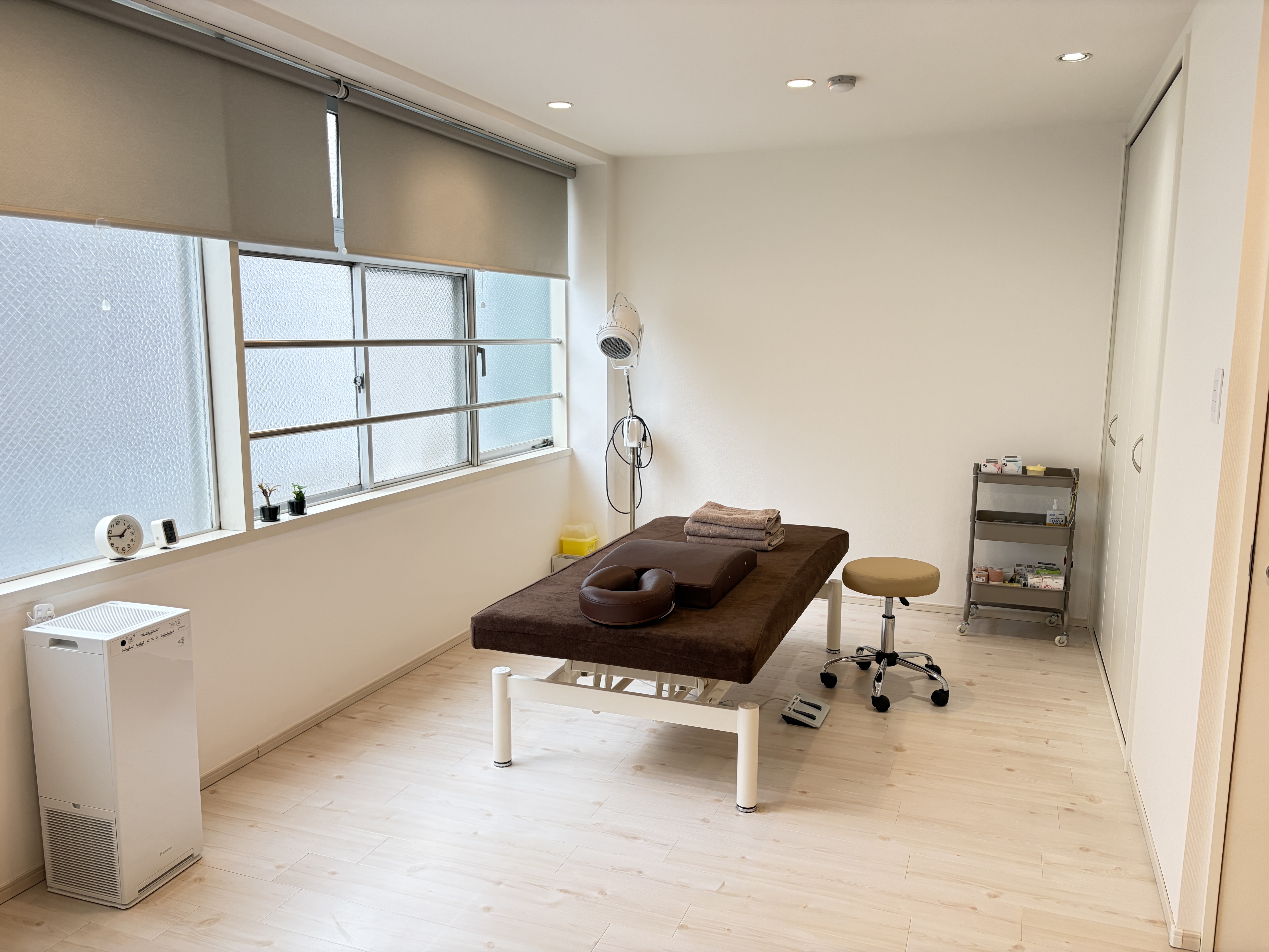 A spa treatment room with a massage table, stool, shelving, and large windows